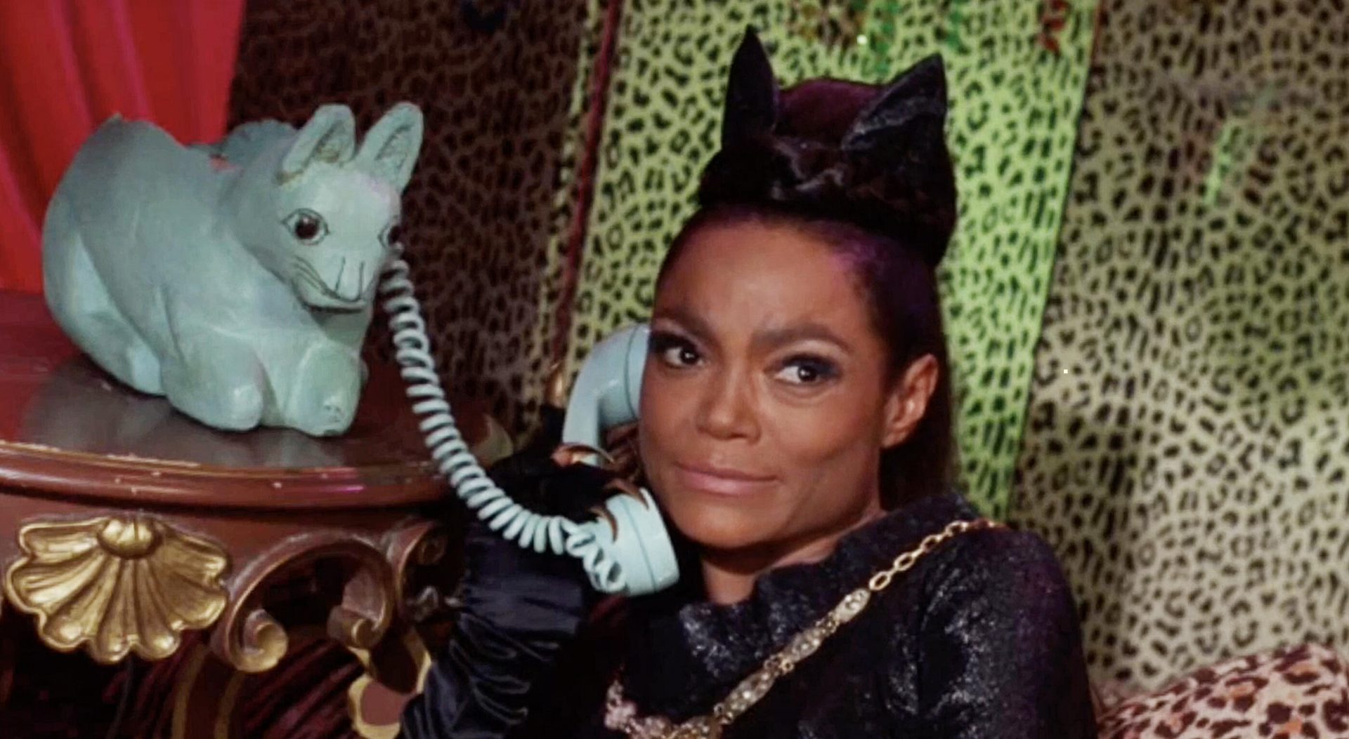 A still from Batman 66 shows Eartha Kitt as Catwoman wearing a black catsuit with a cat on her head speaking into a sheep shaped phone