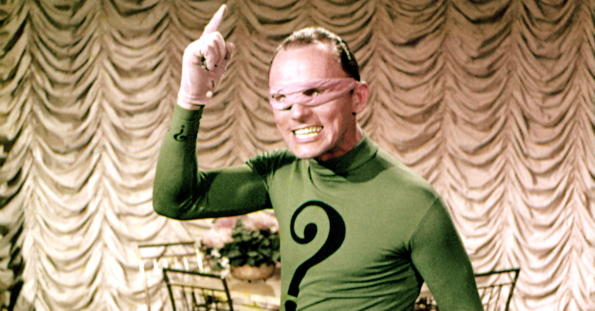 A still from Batman '66 shows Frank Gorshin as the Riddler wearing a pink domino mask and green suit with a large black question mark on it raising his hand menicingly