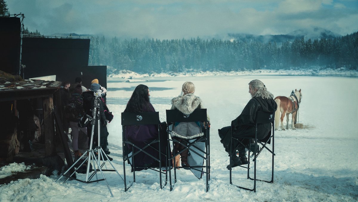 Henry Cavill, Freya Allan, and Anya Chalotra sitting on production chairs near a snowy frozen lake during filming of The Witcher season 3